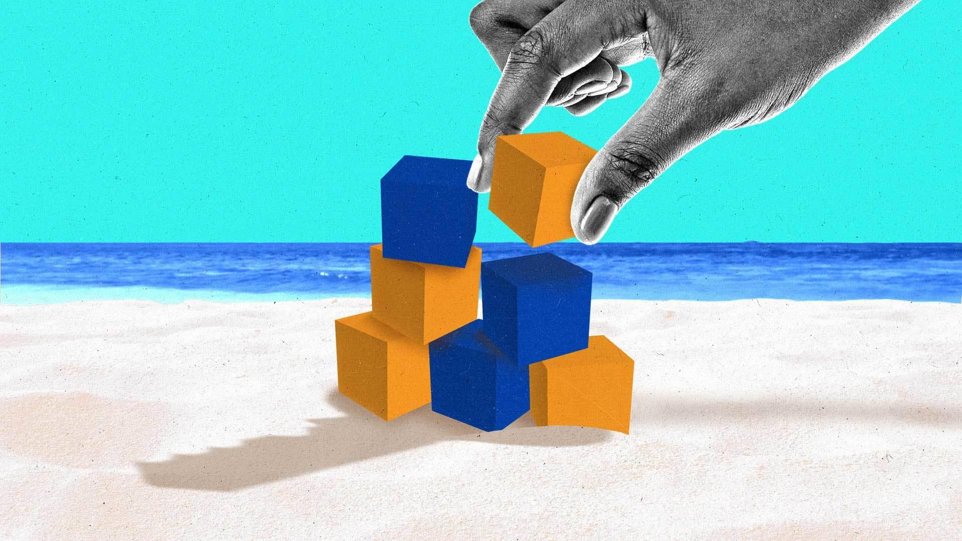 Large hand balances orange and blue boxes on a beach.